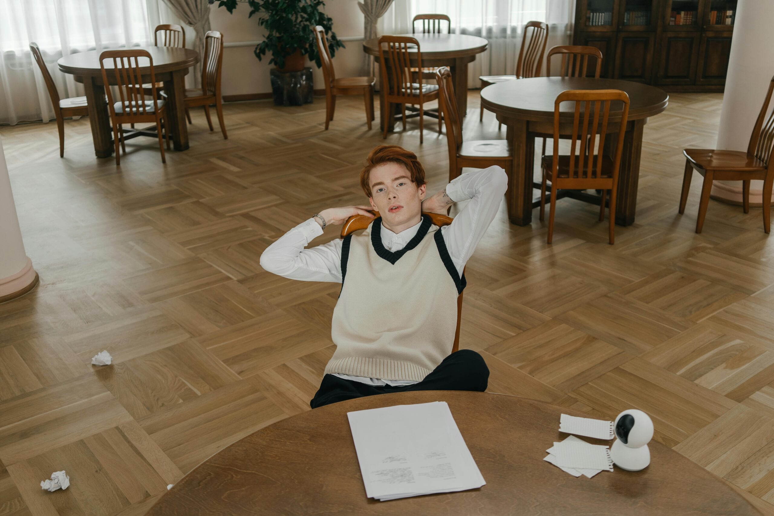 A student in white shirt and vest sits casually in a spacious, wooden-floored study room.
